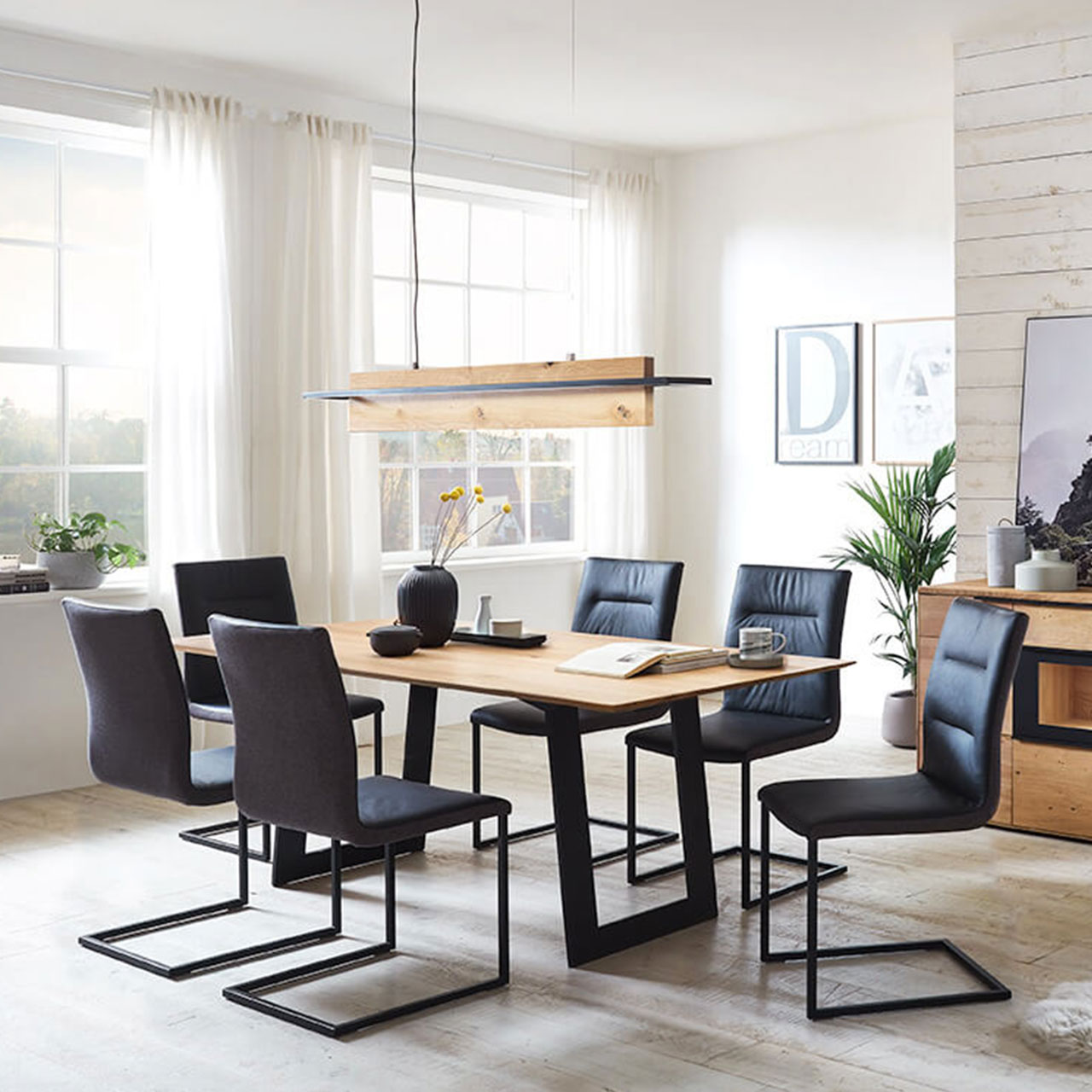Esszimmer-Essgruppe-Ambiente3 Moderner Esstisch mit 6 schwarzen Stühlen als Swinger, dadrüber eine Pendelleuchte in gleichen Wildeiche wie der Tisch und ein schwarzen Metallrahmen.